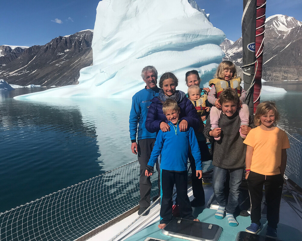 Familie Schw&ouml;rer poseert samen op het dek van hun TOPtoTOP expeditiezeilboot voor een imposante ijsberg in een Groenlandse fjord &ndash; ouders en zes kinderen op een klimaatbewustzijnsreis