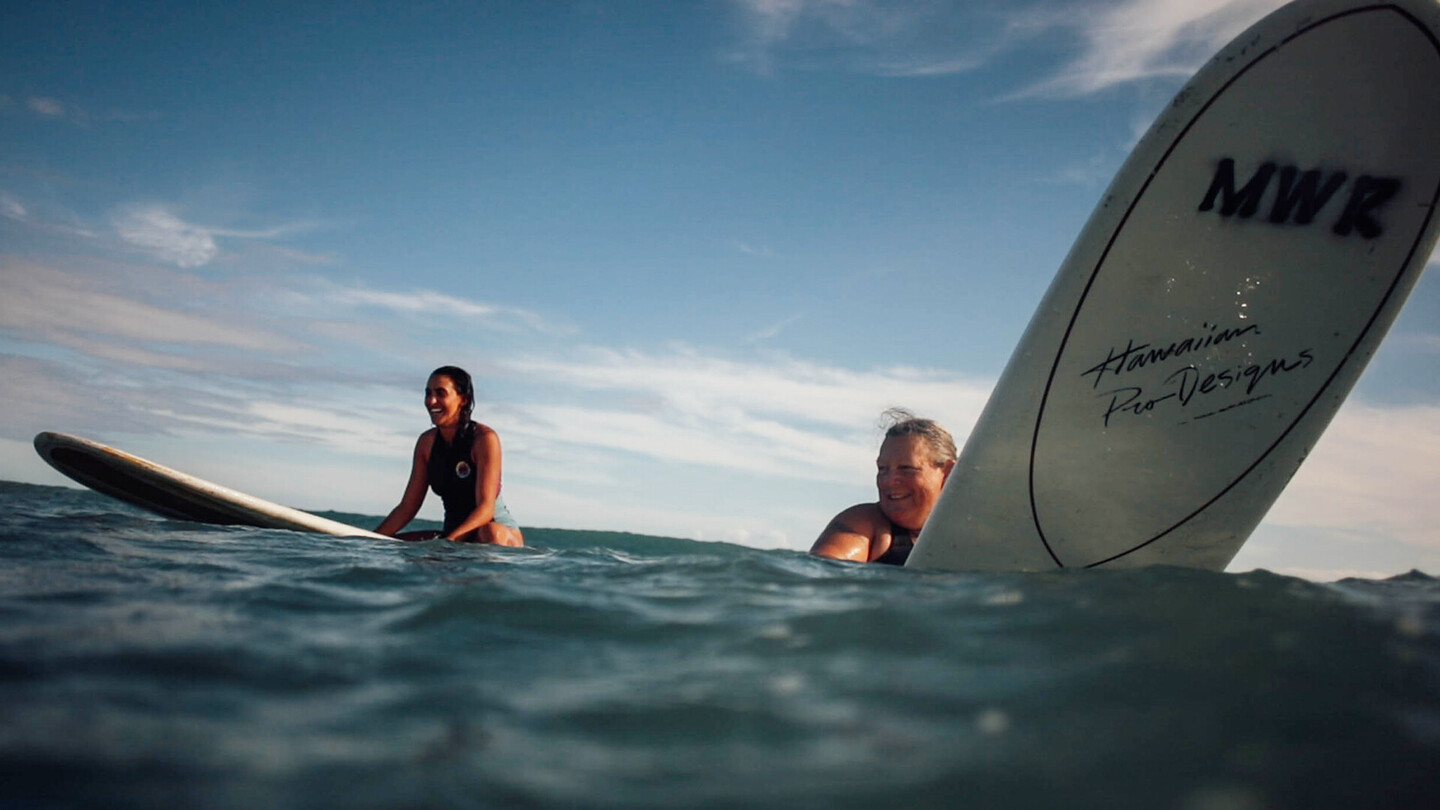 Twee lachende vrouwelijke surfers zitten op hun surfplanken in de open zee, klaar voor de volgende golf.